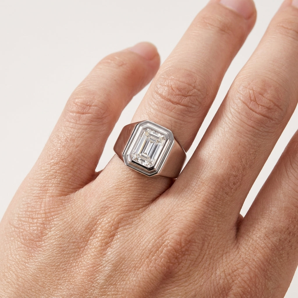 A silver-toned signet ring with an emerald-cut clear gemstone, shown worn on a person's finger against a light off-white background.