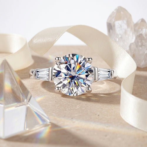 Round moissanite three-stone engagement ring displayed with cream ribbon and crystals