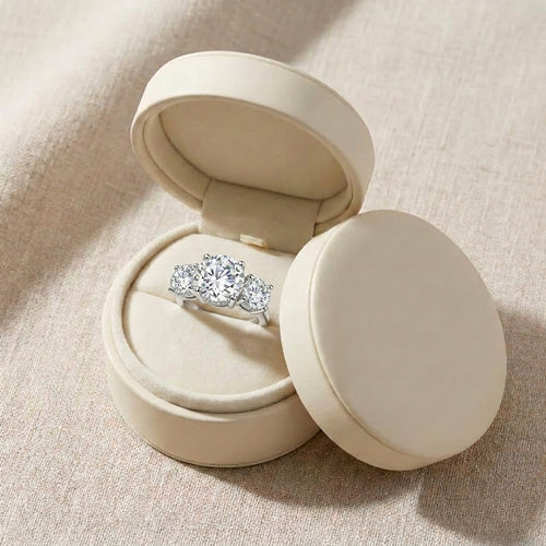 Three-stone round moissanite engagement ring displayed in a cream jewelry box