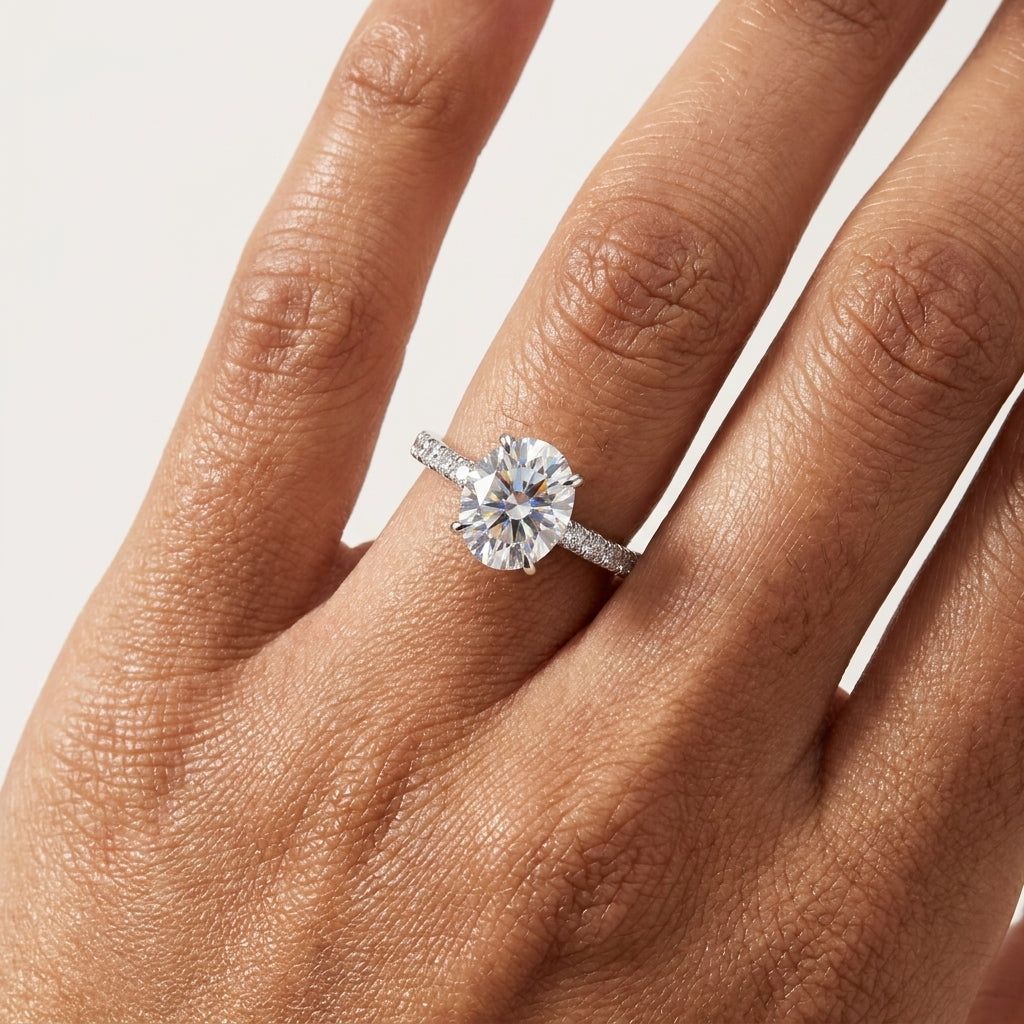 A close-up of a hand wearing a silver-toned engagement ring with a round-cut clear center stone and small side stones on a plain light background.